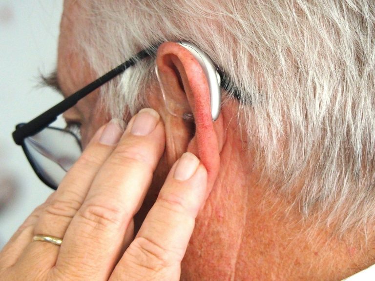 Close up of an older man wearing a hearing aid and glasses viewed from behind his left shoulder. He has his hand on his ear.